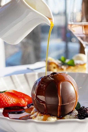 pouring caramel sauce over a chocolate sphere dessert on a white plate